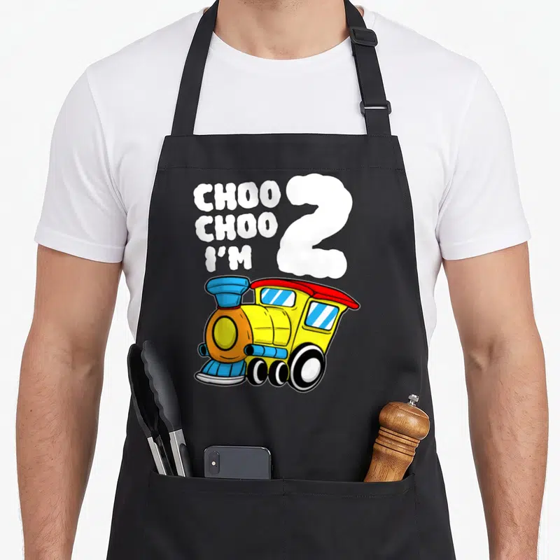 Train 2nd apron - Choo Choo I'm 2 cloud text above yellow blue red cartoon train — shown on a black cotton canvas apron against a white background