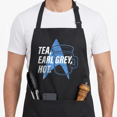 Generation tea apron - Stacked TEA EARL GREY HOT text over blue star and cup outline — shown on a black cotton canvas apron against a white background