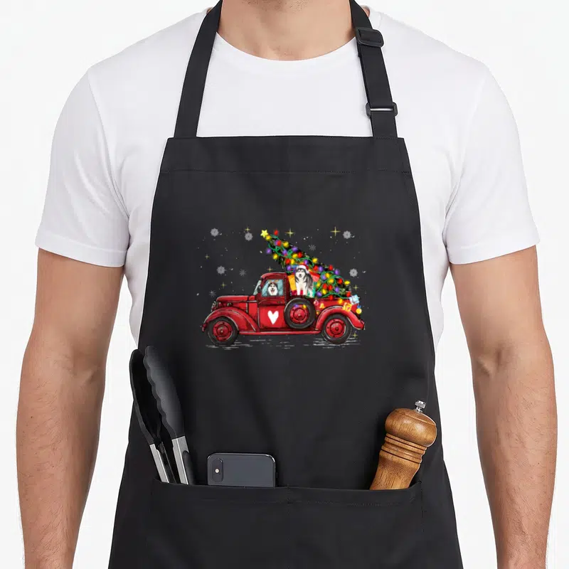 Siberian red christmas apron - Red truck carrying two huskies and a Christmas tree with snowflakes and stars — shown on a black cotton canvas apron against a white background