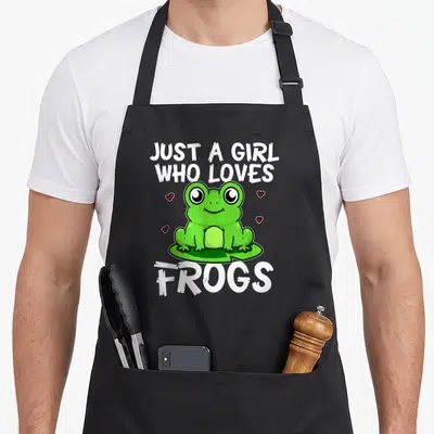 Costume frog aprons - Green cartoon frog on lily pad surrounded by four red hearts with white text Just A Girl Who — shown on a black cotton canvas apron against a white background