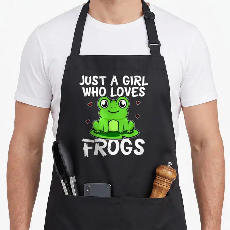 Costume frog aprons - Green cartoon frog on lily pad surrounded by four red hearts with white text Just A Girl Who — shown on a black cotton canvas apron against a white background