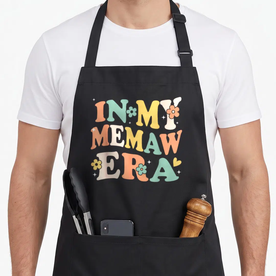 Sarcastic mother's day aprons - Retro text In My Memaw Era with flowers and stars in orange green yellow white — shown on a black cotton canvas apron against a white background