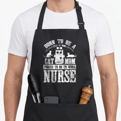 Forced at home aprons - White distressed text BORN TO BE A STAY AT HOME CAT MOM FORCED TO GO TO WORK NURSE surrounding — shown on a black cotton canvas apron against a white background