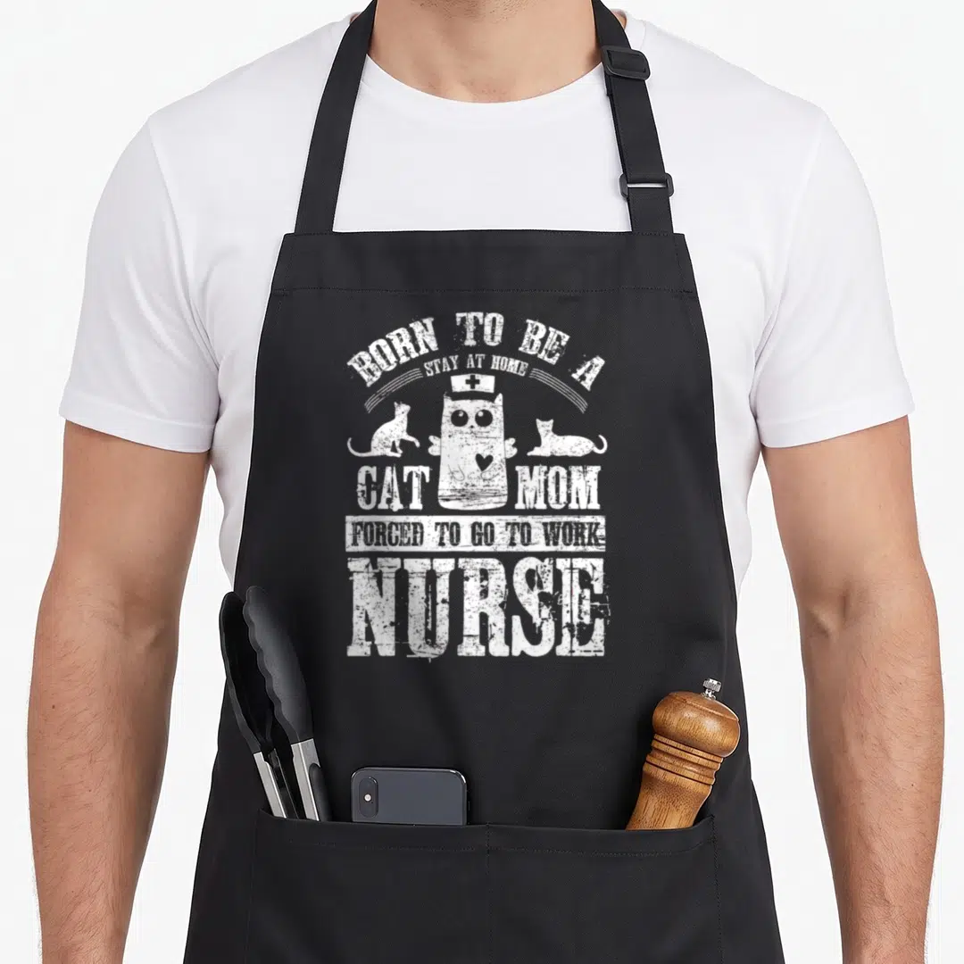 Forced at home aprons - White distressed text BORN TO BE A STAY AT HOME CAT MOM FORCED TO GO TO WORK NURSE surrounding — shown on a black cotton canvas apron against a white background