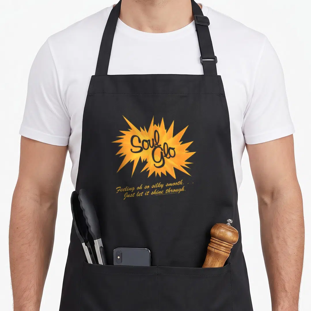 Coming soul apron - Orange starburst with grey Soul Glo text and yellow cursive tagline on grey background — shown on a black cotton canvas apron against a white background