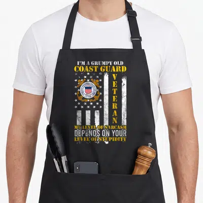 Grumpy coast guard apron - Distressed flag background with centered Coast Guard emblem and vertical veteran text — shown on a black cotton canvas apron against a white background
