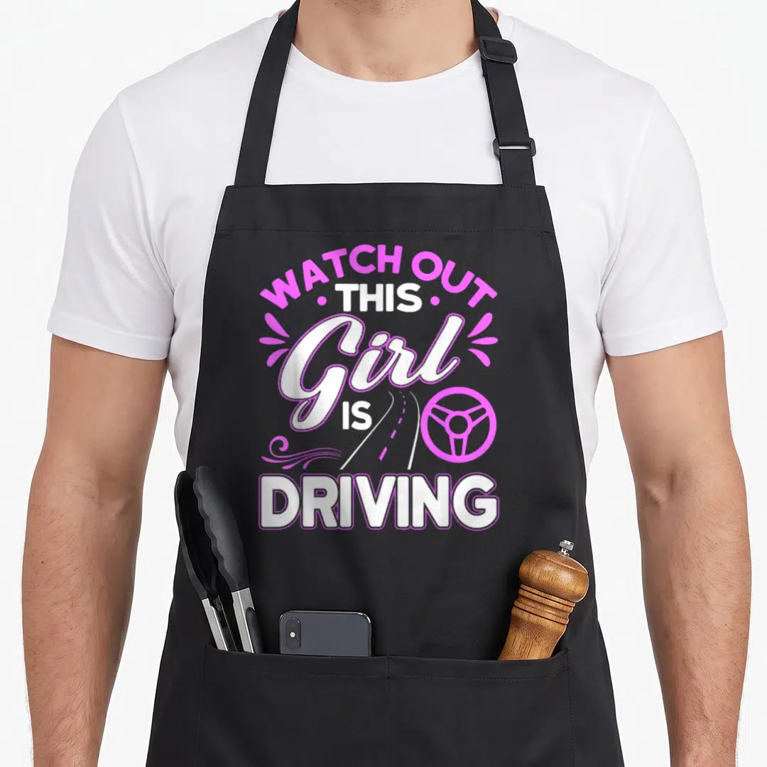 Passed driving apron - Pink and white text reading WATCH OUT THIS Girl IS DRIVING with steering wheel icon — shown on a black cotton canvas apron against a white background