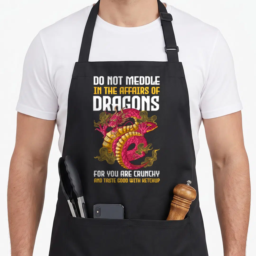 Affair dragon apron - Pink cartoon dragon surrounded by text DO NOT MEDDLE IN THE AFFAIRS OF DRAGONS FOR YOU ARE — shown on a black cotton canvas apron against a white background