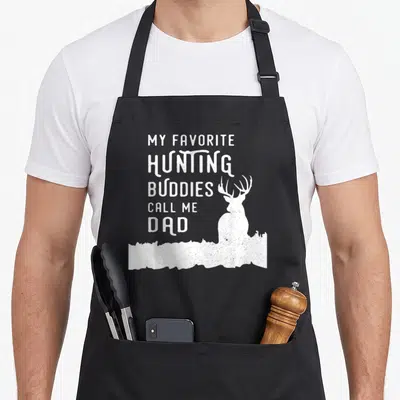 Hunter dad and son aprons - White distressed text My Favorite Hunting Buddies Call Me Dad with deer silhouette in grass — shown on a black cotton canvas apron against a white background
