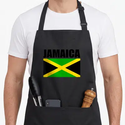 Cute flag jamaican apron - JAMAICA text above rectangular flag with black green and yellow cross pattern — shown on a black cotton canvas apron against a white background