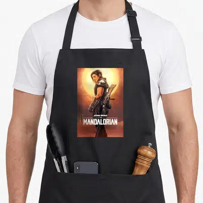 Cara little miss apron - Cara Dune in armor standing before a sun with STAR WARS THE MANDALORIAN text at the bottom — shown on a black cotton canvas apron against a white background