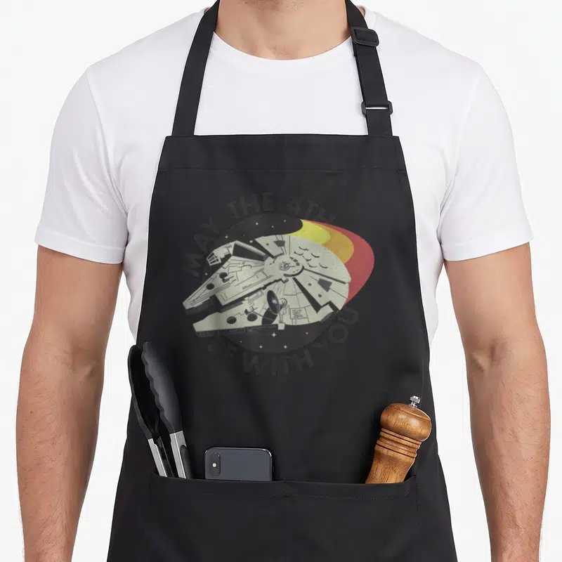 4th vintage may apron - Retro Millennium Falcon with MAY THE 4TH text and yellow orange red semi-circles — shown on a black cotton canvas apron against a white background