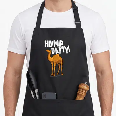 Camel dromedary apron - Brown cartoon camel standing under white HUMP DAYYY! text — shown on a black cotton canvas apron against a white background