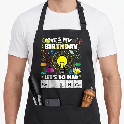 Theme science apron - Light bulb surrounded by flasks, Einstein heads, and periodic table cells spelling ScIENCe — shown on a black cotton canvas apron against a white background