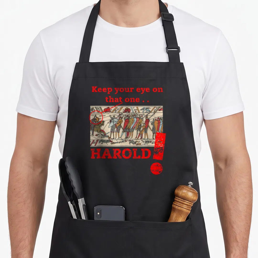 Medieval quirky apron - Distressed red text Keep your eye on that one Harold above Bayeux Tapestry scene with circled — shown on a black cotton canvas apron against a white background