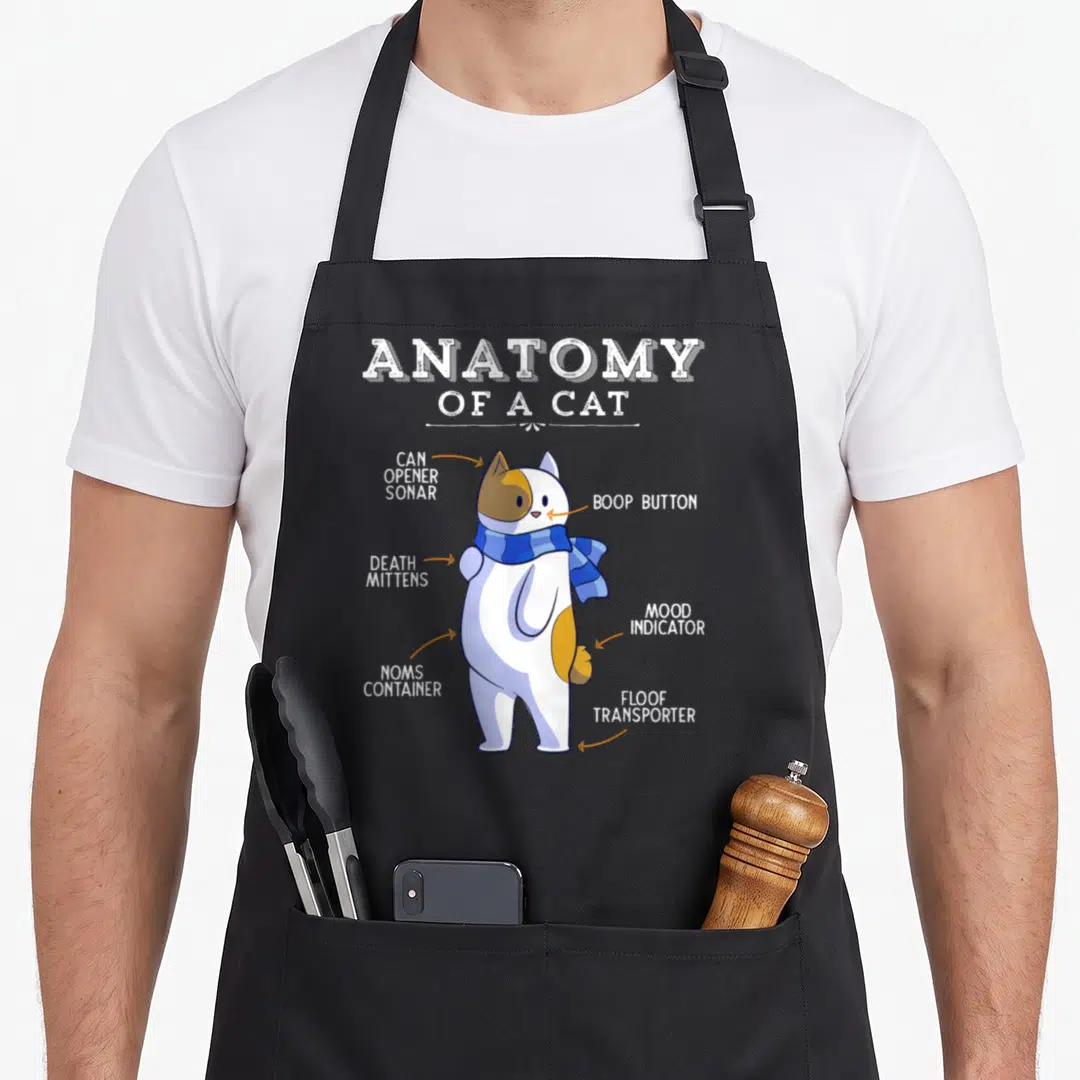 Anatomy kitty cat apron - Cartoon cat diagram with labels Boop Button, Noms Container, and Floof Transporter in blue — shown on a black cotton canvas apron against a white background