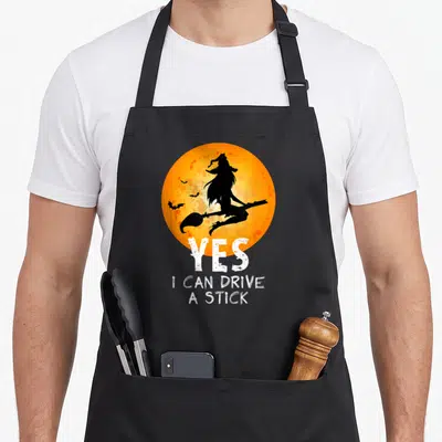Funny drive halloween witch apron - Witch silhouette on orange moon with Yes I Can Drive A Stick text — shown on a black cotton canvas apron against a white background
