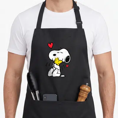 Woodstock lot valentine apron - White cartoon dog hugging yellow bird with two red hearts — shown on a black cotton canvas apron against a white background