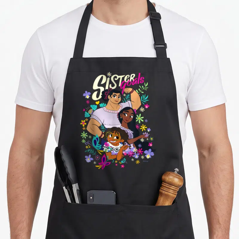 Mirabel sister goals apron - Three sisters stacked vertically under arched Sister goals text with purple, yellow, pink — shown on a black cotton canvas apron against a white background