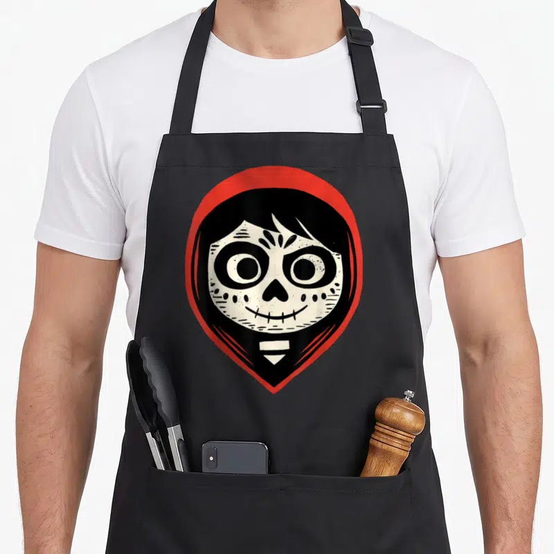 Coco sugar skull apron - White cartoon skull face inside black shape bordered by red shield outline — shown on a black cotton canvas apron against a white background