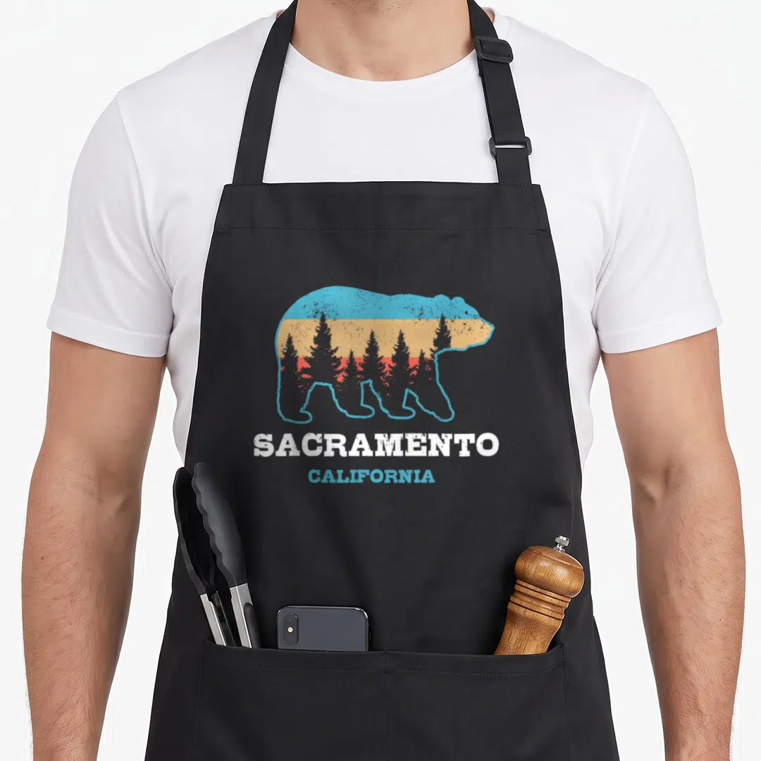 Sacramento bear apron - Grizzly bear silhouette with striped trees and Sacramento California text — shown on a black cotton canvas apron against a white background