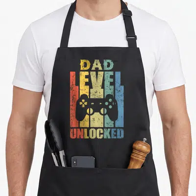 Level pregnancy announcement apron - Retro DAD LEVEL UNLOCKED text with grey game controller and rainbow stripes — shown on a black cotton canvas apron against a white background