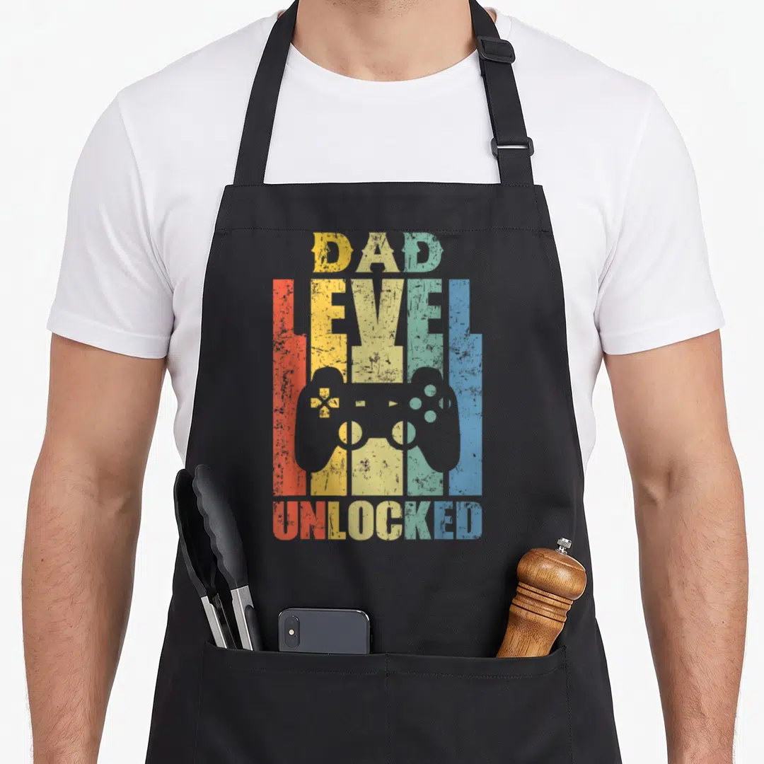 Level pregnancy announcement apron - Retro DAD LEVEL UNLOCKED text with grey game controller and rainbow stripes — shown on a black cotton canvas apron against a white background