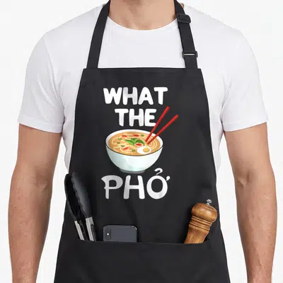 Noodle bowl pho apron - Cartoon bowl of pho with red chopsticks and text WHAT THE PHỞ in white — shown on a black cotton canvas apron against a white background