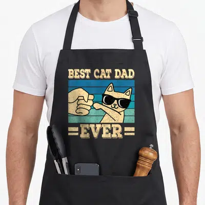Cute cat dad apron - Cartoon cat with sunglasses fist-bumping human hand under Best Cat Dad Ever text — shown on a black cotton canvas apron against a white background