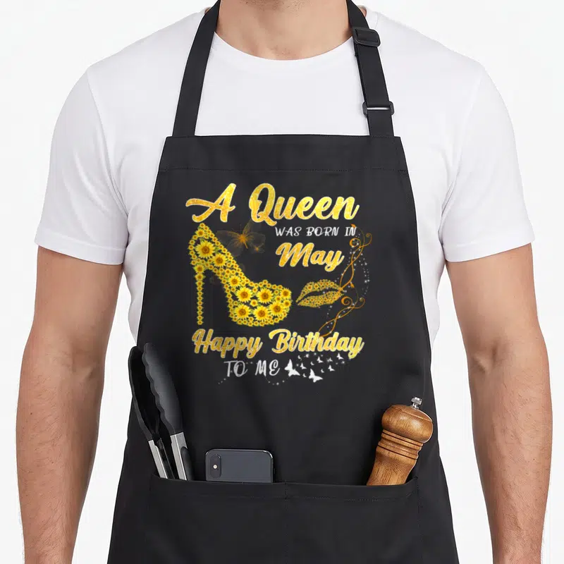 Queen sunflower apron - High heel shoe made of sunflowers with gold text A Queen Was Born In May Happy Birthday To Me — shown on a black cotton canvas apron against a white background