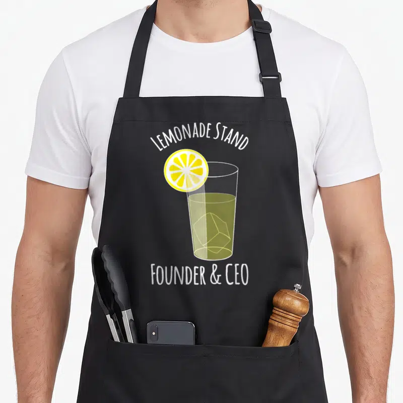 Entrepreneur lemonade apron - Cartoon glass with yellow slice and white text LEMONADE STAND FOUNDER CEO — shown on a black cotton canvas apron against a white background
