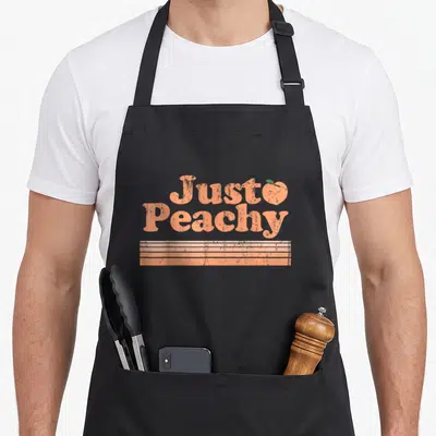 Peach fruit apron - Just Peachy retro text with peach icon and three horizontal stripes — shown on a black cotton canvas apron against a white background