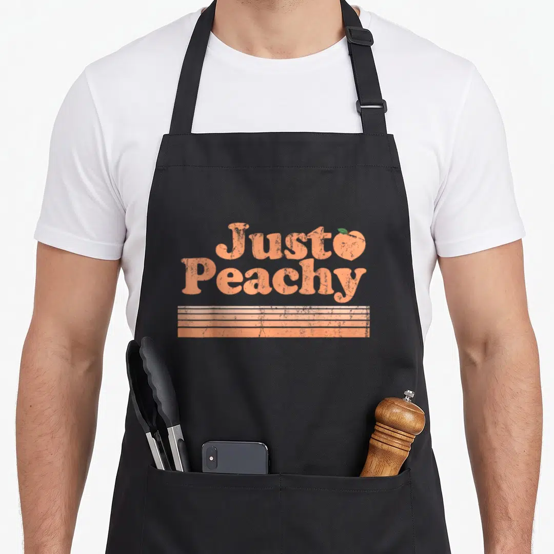 Peach fruit apron - Just Peachy retro text with peach icon and three horizontal stripes — shown on a black cotton canvas apron against a white background