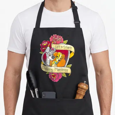 Tramp building apron - Two dogs inside heart frame with cream ribbon text and red roses — shown on a black cotton canvas apron against a white background