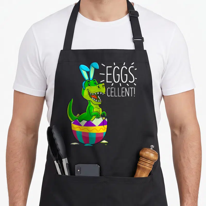 Stuffer basket apron - Green dinosaur with blue bunny ears hatching from colorful egg beside white EGGS CELLENT text — shown on a black cotton canvas apron against a white background