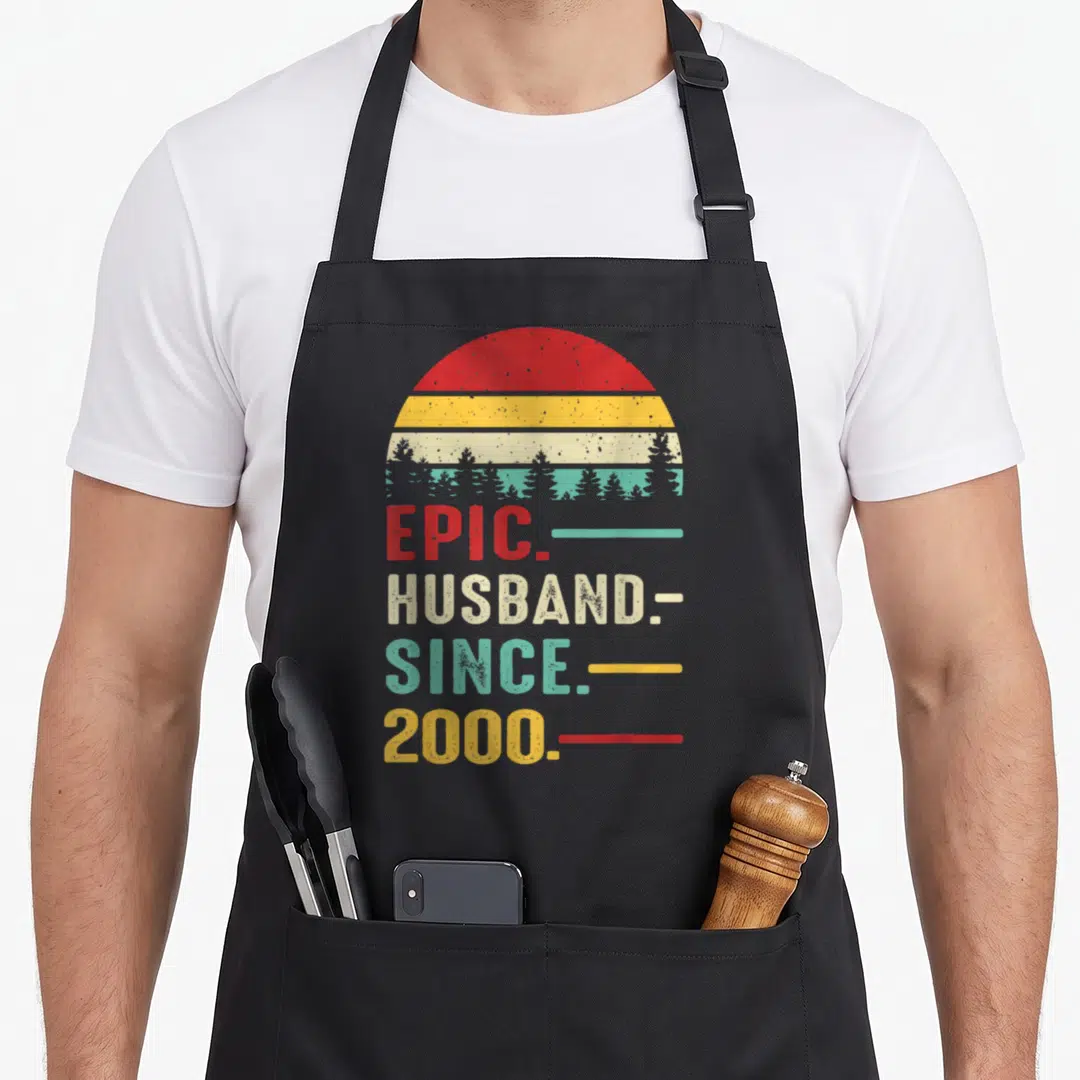 26th wedding aprons - Retro sunset with tree silhouettes above stacked text reading epic husband since 2000 in red — shown on a black cotton canvas apron against a white background