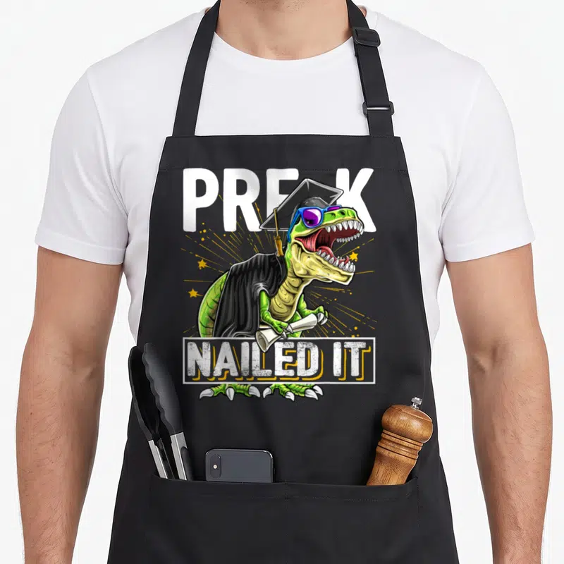 Graduation dinosaur apron - Tyrannosaurus Rex in cap gown holding diploma with PRE-K Nailed It text — shown on a black cotton canvas apron against a white background