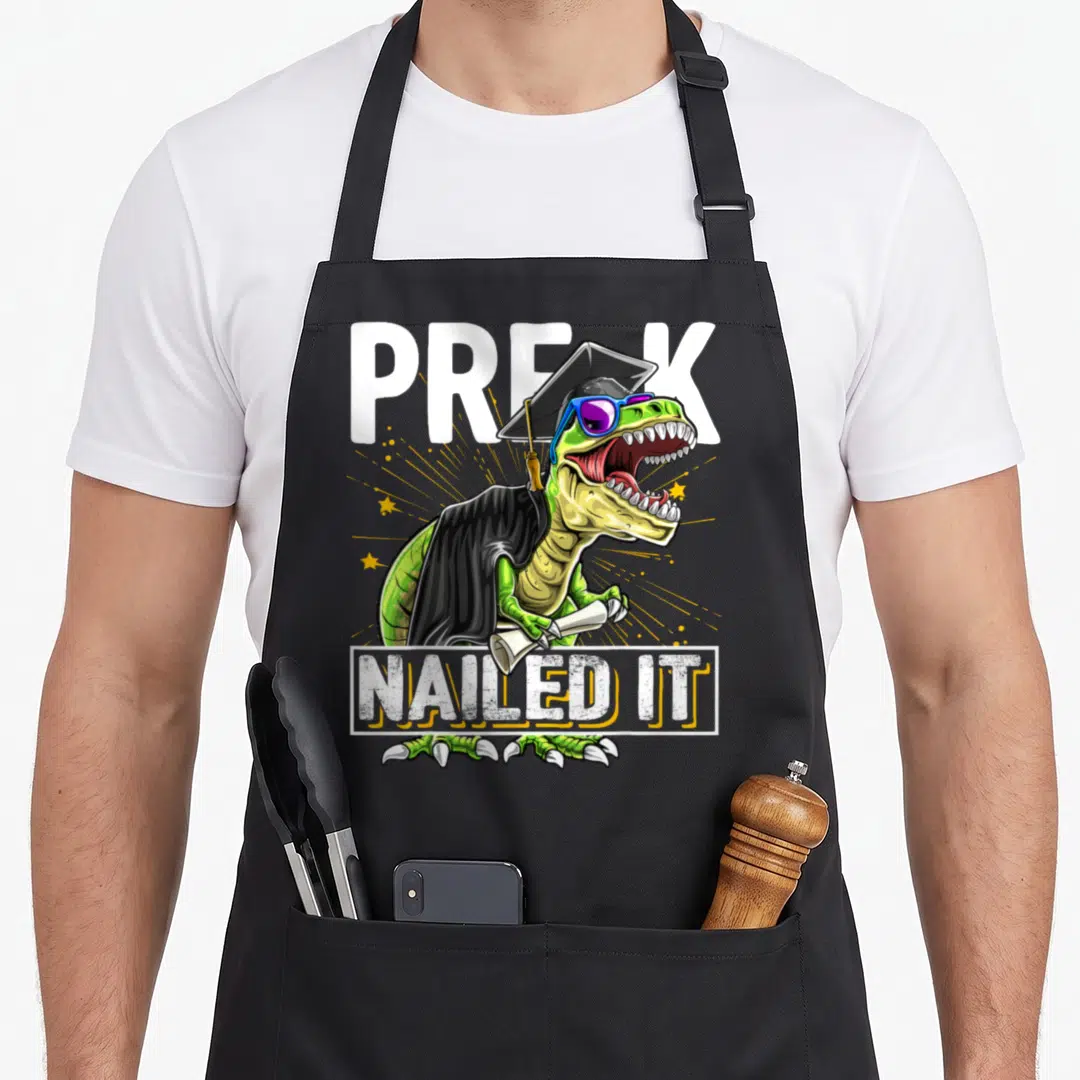 Graduation dinosaur apron - Tyrannosaurus Rex in cap gown holding diploma with PRE-K Nailed It text — shown on a black cotton canvas apron against a white background