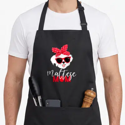 Maltese fur dog apron - Cartoon Maltese head with red sunglasses and polka dot bow above text — shown on a black cotton canvas apron against a white background