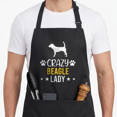 Funny beagle dog cooking apron - White beagle silhouette in grey halftone circle with stacked text and paw prints — shown on a black cotton canvas apron against a white background