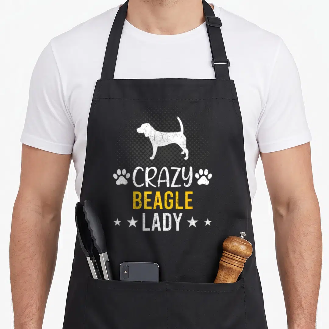 Funny beagle dog cooking apron - White beagle silhouette in grey halftone circle with stacked text and paw prints — shown on a black cotton canvas apron against a white background