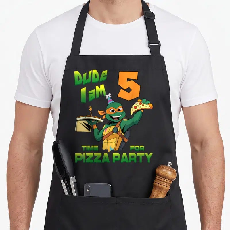Dude mikey pizza apron - Cartoon turtle holding pizza box with candle and slice surrounded by birthday text — shown on a black cotton canvas apron against a white background