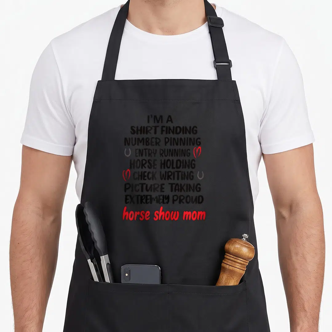 Show definition mother's apron - Stacked black text listing equestrian duties with red horse show mom and grey — shown on a black cotton canvas apron against a white background