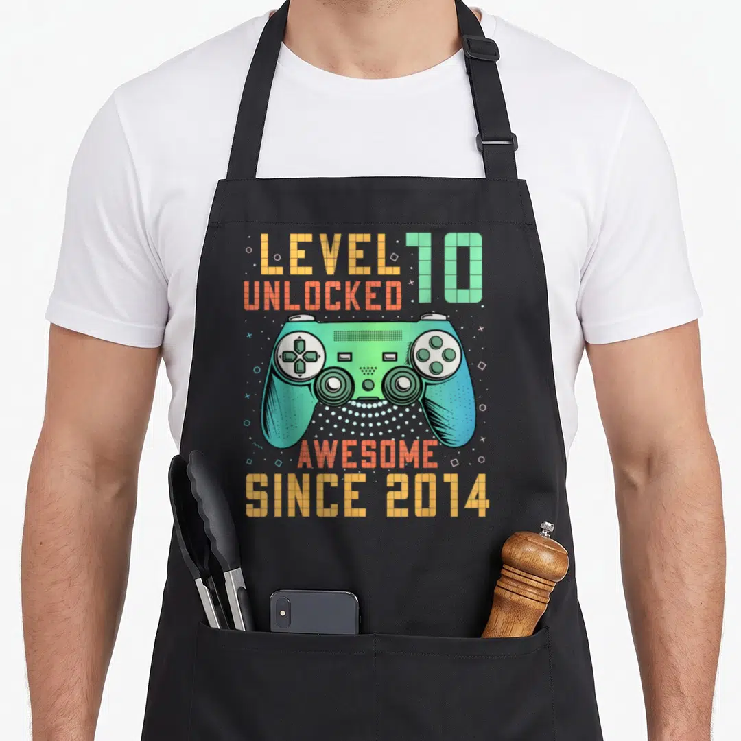 Unlocked 10th birthday apron - Pixel art game controller with Level 10 Unlocked and Awesome Since 2014 text in orange — shown on a black cotton canvas apron against a white background