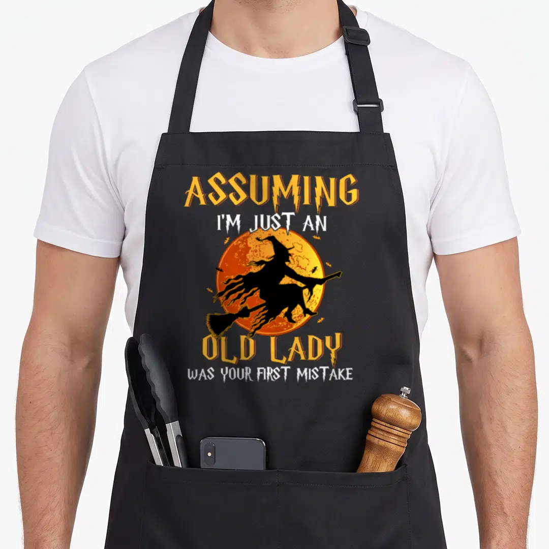 Assuming witch apron - Cartoon witch silhouette on broom against orange moon with sarcastic text about being an old lady — shown on a black cotton canvas apron against a white background