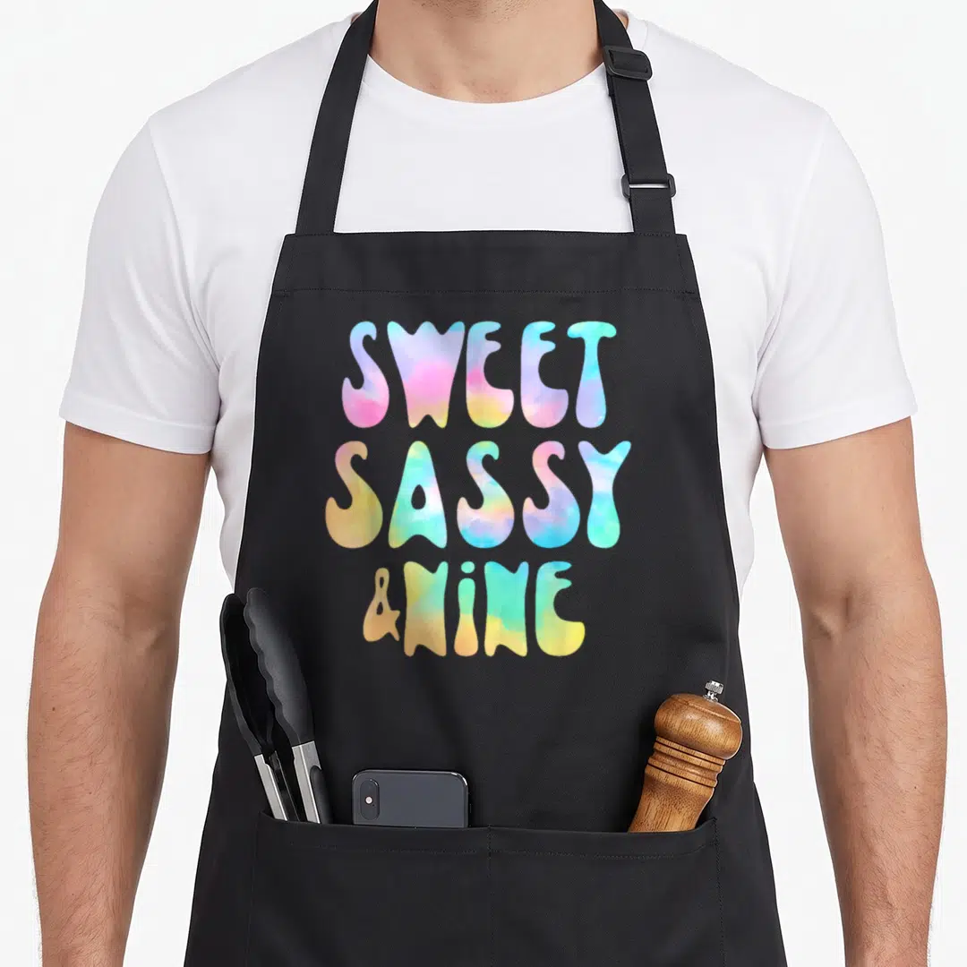 Nine sassy apron - Stacked text reading Sweet Sassy and Nine in pink blue yellow and green tie dye font — shown on a black cotton canvas apron against a white background