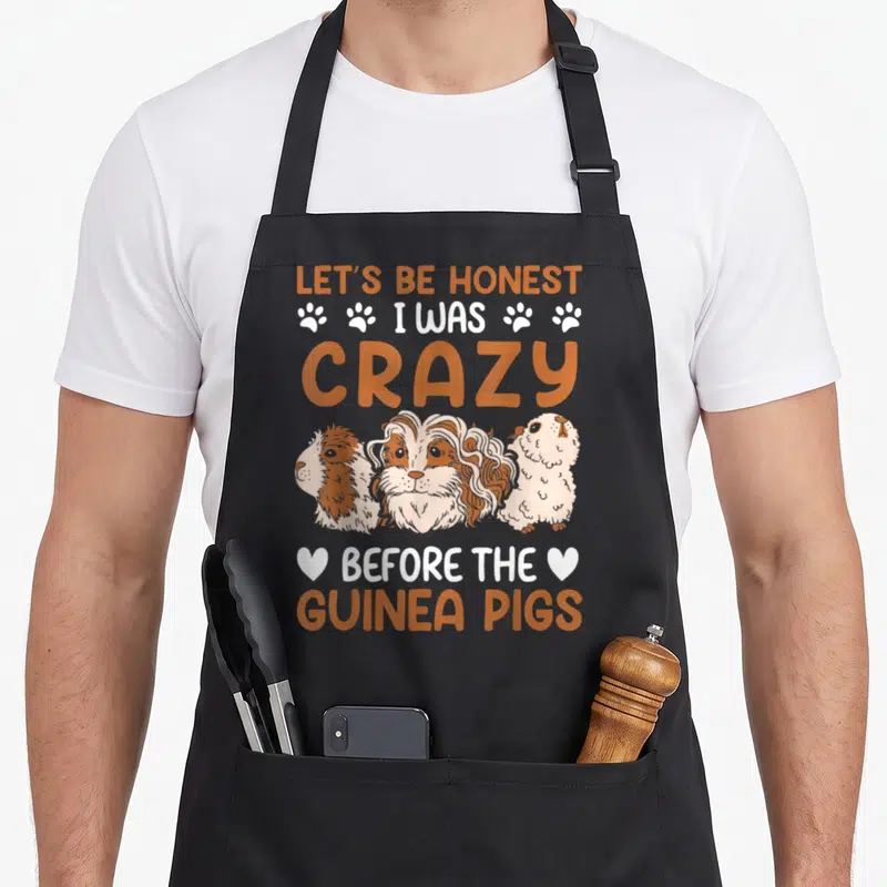 Before crazy guinea man apron - Three cartoon guinea pigs with orange and white text and paw prints — shown on a black cotton canvas apron against a white background