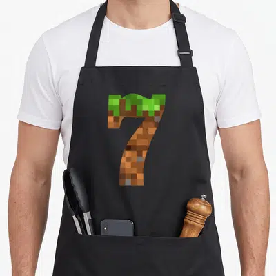Pixel 7yr birthday apron - Pixelated number 7 formed by green grass and brown dirt blocks — shown on a black cotton canvas apron against a white background