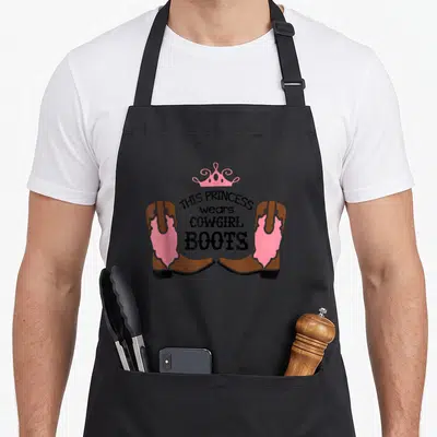 Boot western apron - Two brown and pink cowgirl boots flanking black text under a pink crown — shown on a black cotton canvas apron against a white background