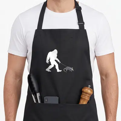 Armadillo walking apron - White Bigfoot silhouette walking right holding a leash attached to a small armadillo — shown on a black cotton canvas apron against a white background
